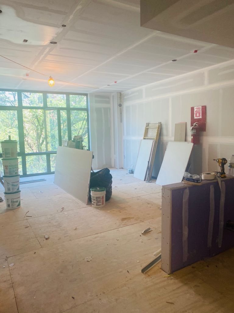 Interior of a room under construction with drywall and plywood flooring, large window, construction tools, and supplies.