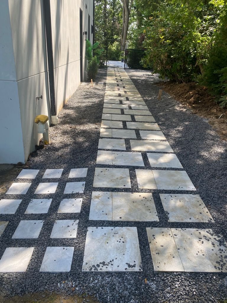 A newly paved walkway with white tiles set in black gravel, running alongside a building with a white exterior, bordered by green trees and plants on one side and a gravel path on the other.