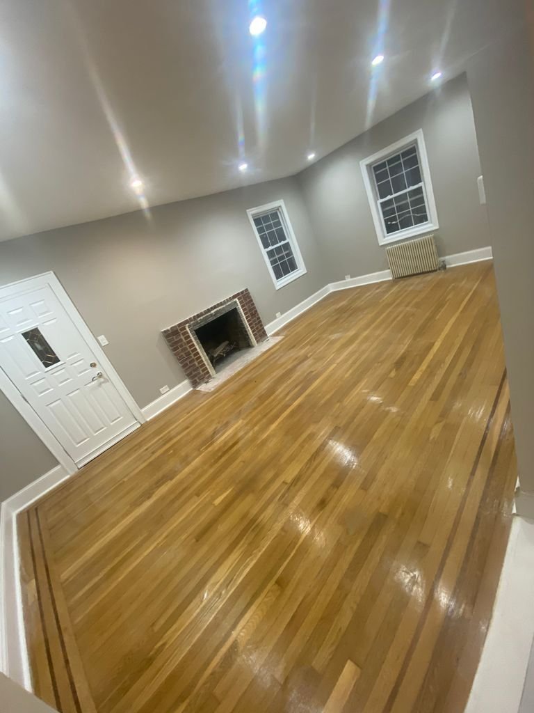 Empty living room with hardwood floors, gray walls, two windows, a brick fireplace, white door, and an overhead light reflected on the floor.