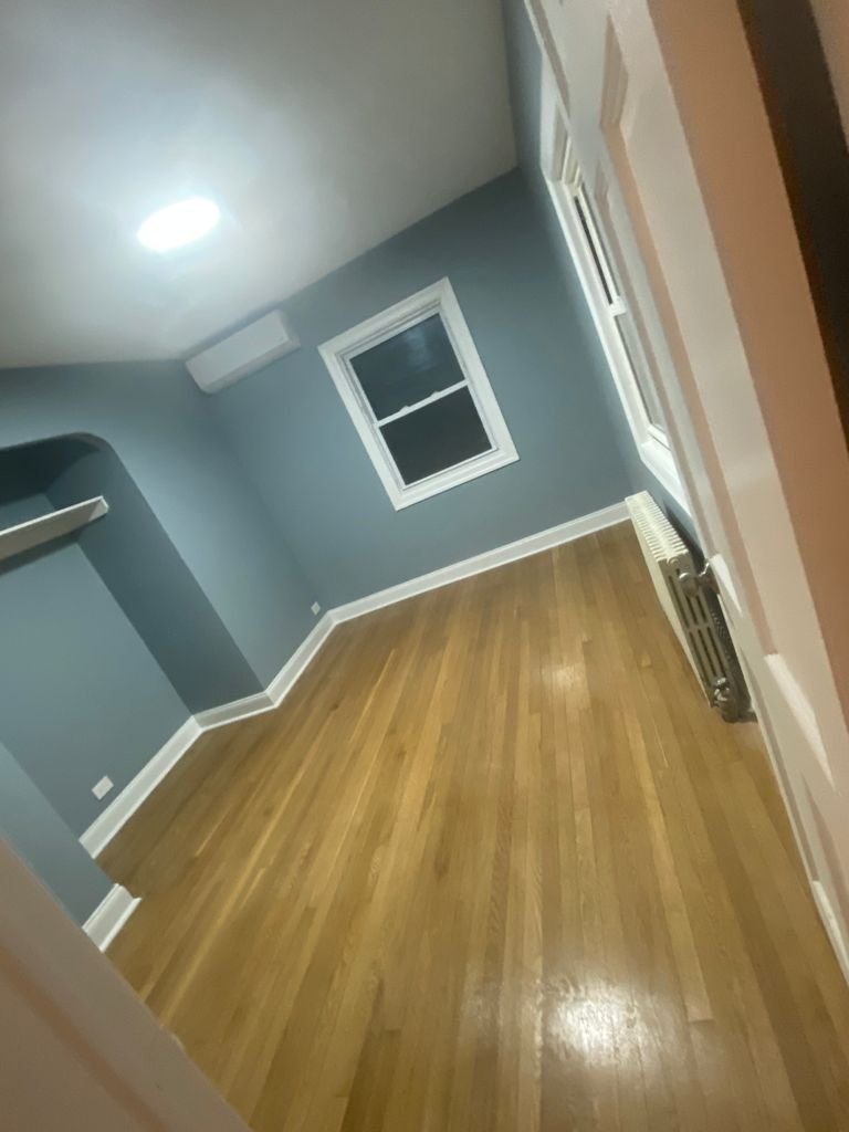 Empty room with hardwood floor, blue walls, two windows, a white radiator, and a ceiling light.