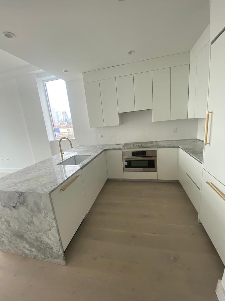Modern kitchen with white cabinets, marble countertops, built-in oven, and a sink near a large window with city view.
