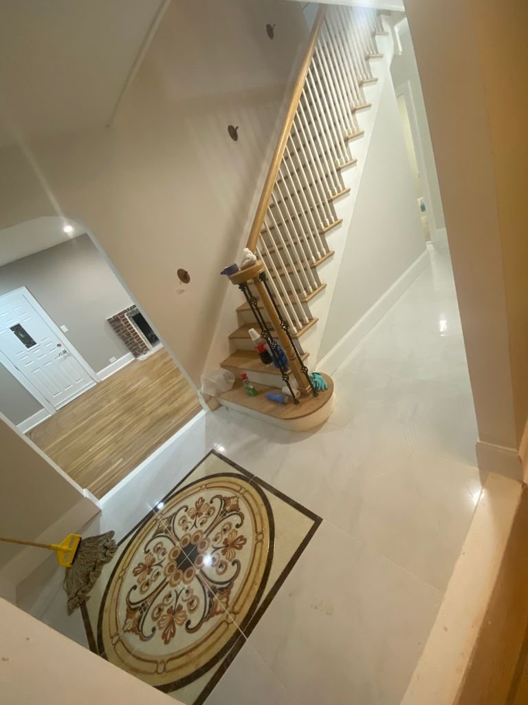 Entrance foyer with a decorative floor tile, staircase with wooden handrail, and cleaning supplies on the floor.