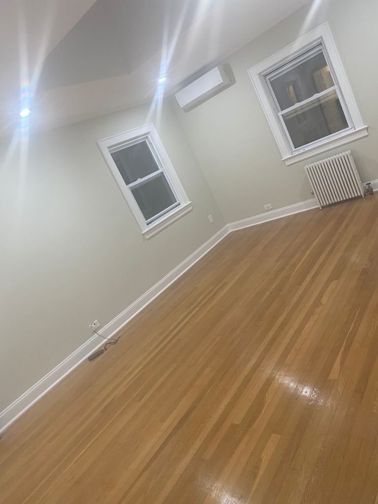 Empty room with hardwood floors, two windows, white walls, an air conditioner, baseboards, radiator, and an electrical outlet.