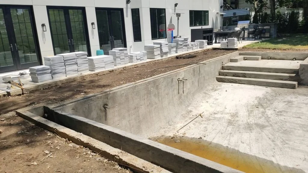 Empty in-ground swimming pool under construction with building in background, construction materials along the side, and stairs at one end.