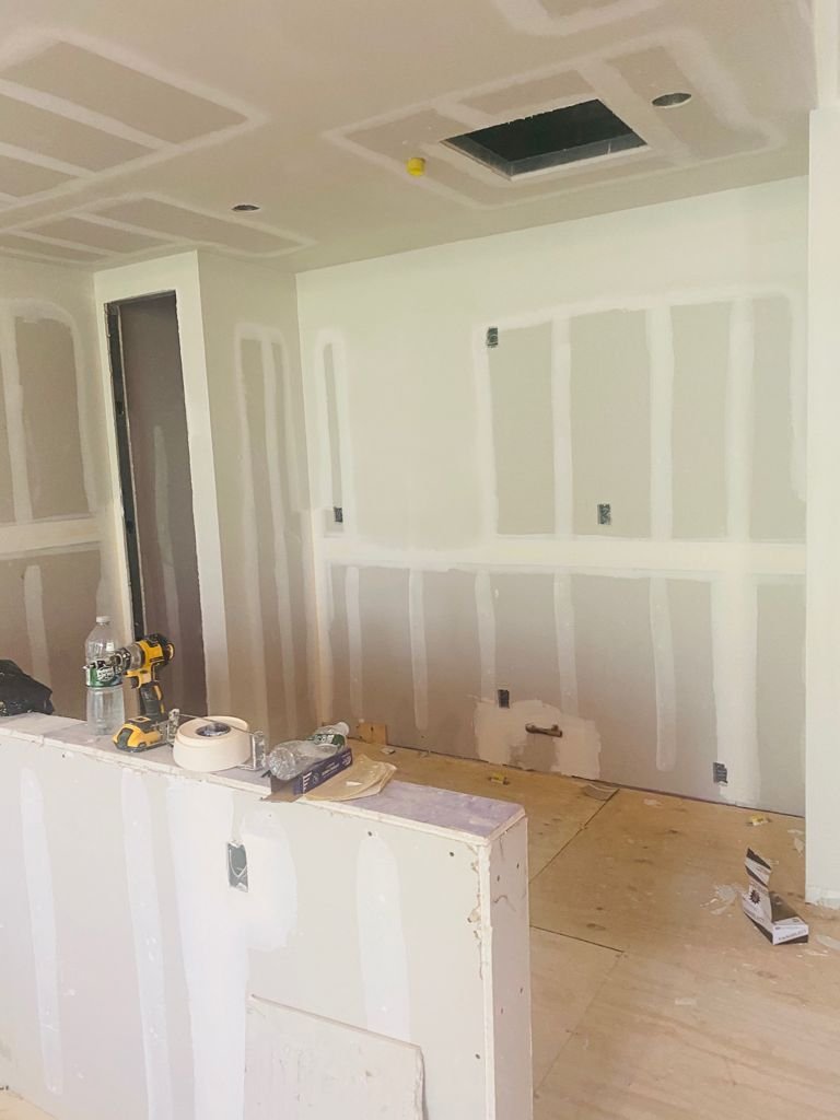 Interior room under construction with drywall on walls and ceiling, electrical outlets and switches installed, construction tools and materials on a partially built counter or wall, and an opening in the floor covered with plywood.