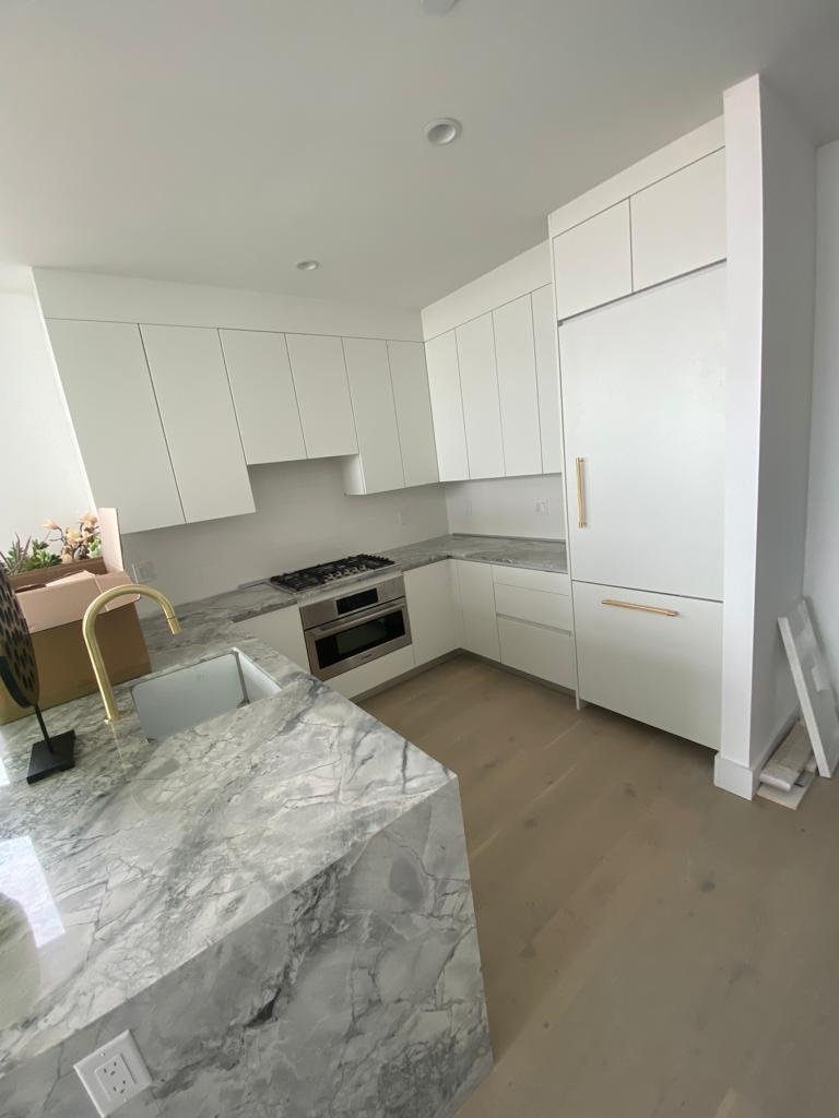 Modern kitchen with white cabinetry, marble countertops, a built-in oven, stove, and a gold-colored faucet over a marble island. The floor is light wood, and there are some items on the countertop and a ladder leaning against the wall.