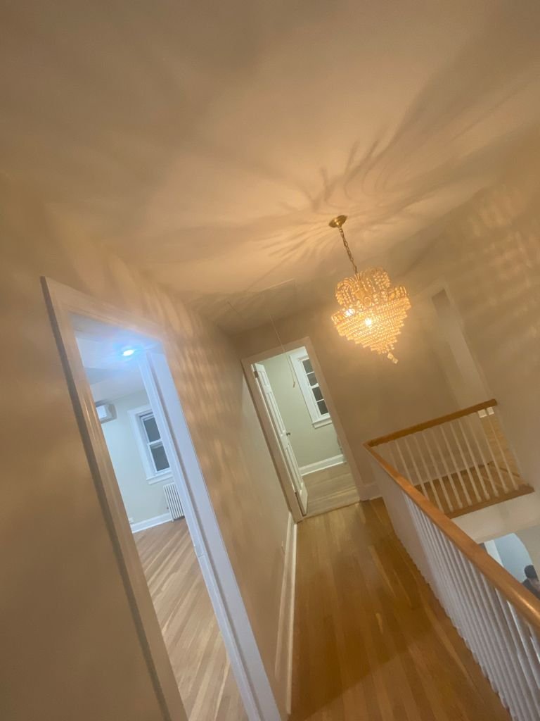 Hallway with chandelier, wooden flooring, and two open doors leading to rooms with windows.