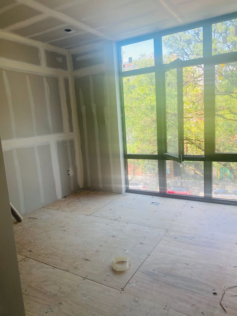 An unfinished room with drywall and a large window showing trees outside, with sunlight streaming in.