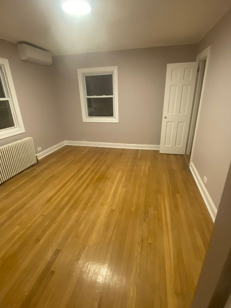 Empty room with hardwood floors, two windows with white trim, a radiator, a white door, and beige walls.