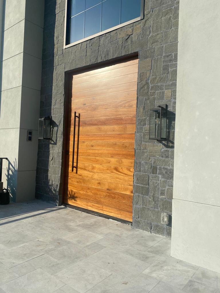 Modern house entrance with large wooden door, black metal handles, stone wall exterior, black wall-mounted lantern, and tiled walkway.