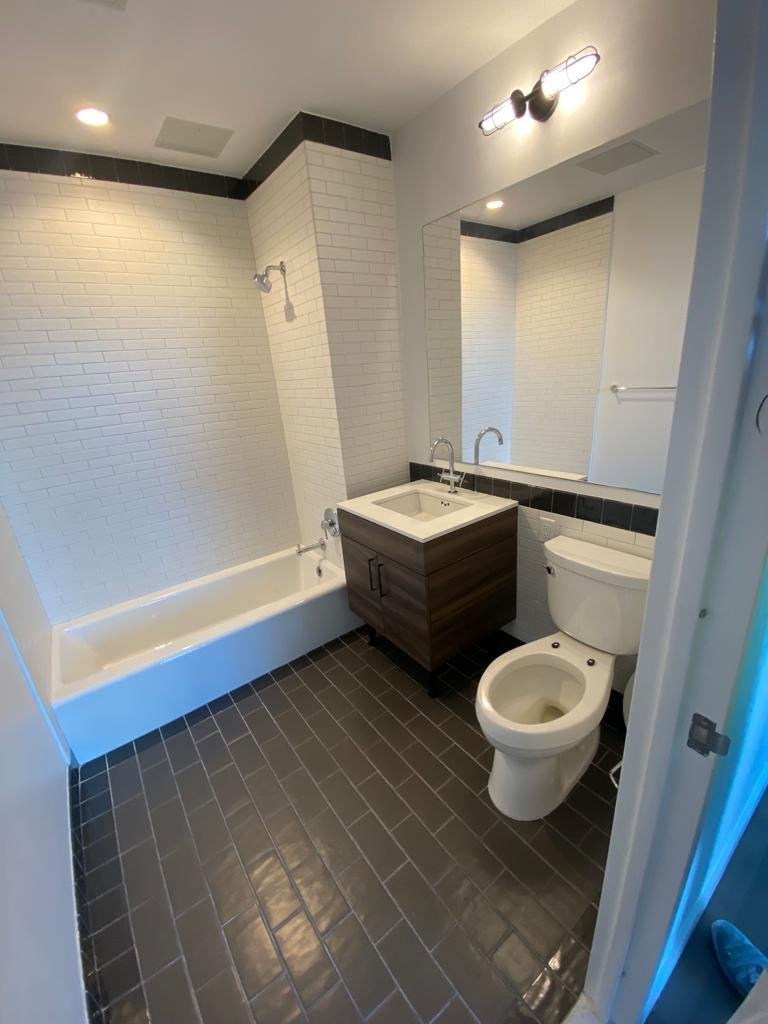 A modern bathroom with a white bathtub, a dark wood vanity with a white sink, a large mirror, a toilet, and a combination shower and bathtub area with white brick walls and black tile flooring.