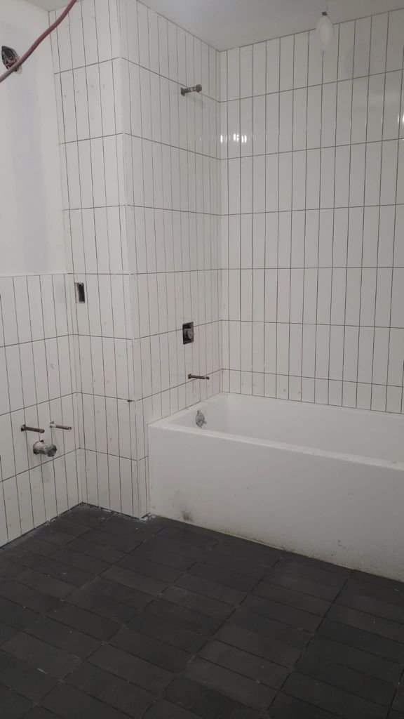 Bathroom with white tiled walls, a bathtub on the right, and plumbing fixtures for a sink or washing machine left of the bathtub. The floor is covered with dark tiles.