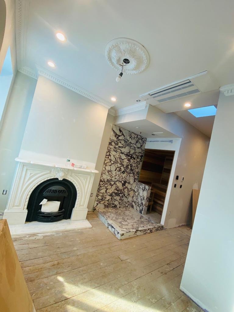 Interior view of a living space under renovation, featuring a decorative fireplace with ornate molding, a newly installed marble section on the wall and floor, and visible light fixtures including a ceiling medallion with a hanging bulb