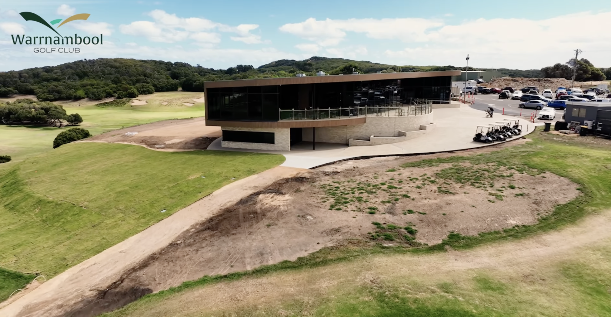 A modern golf club building with large glass windows and a parking lot filled with cars at Warnnambool Golf Club, surrounded by green grass and hills under a partly cloudy sky.