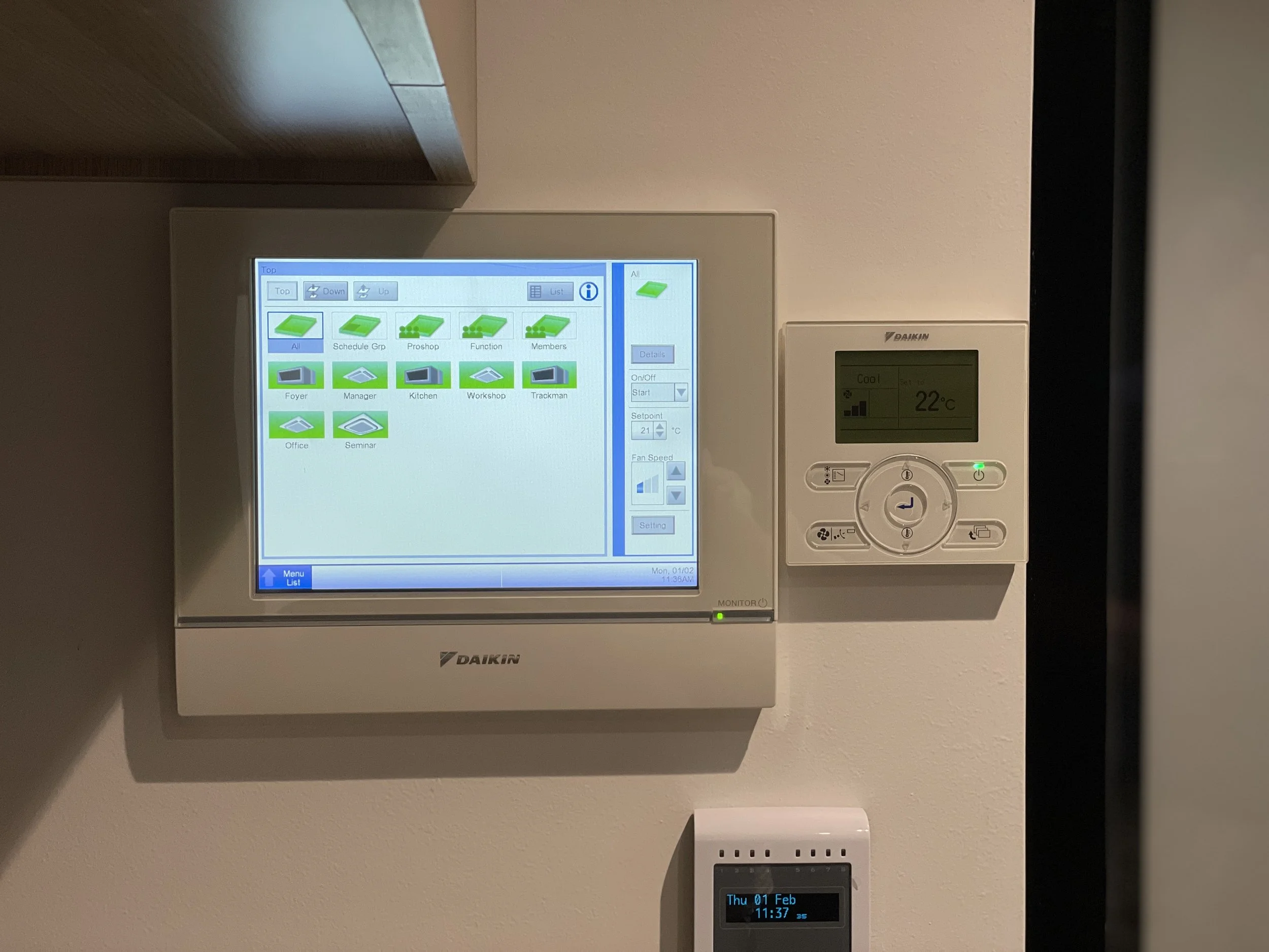 Digital control panel with a touchscreen display showing building zones, beside a smaller thermostat set to 22°C, mounted on a white wall.