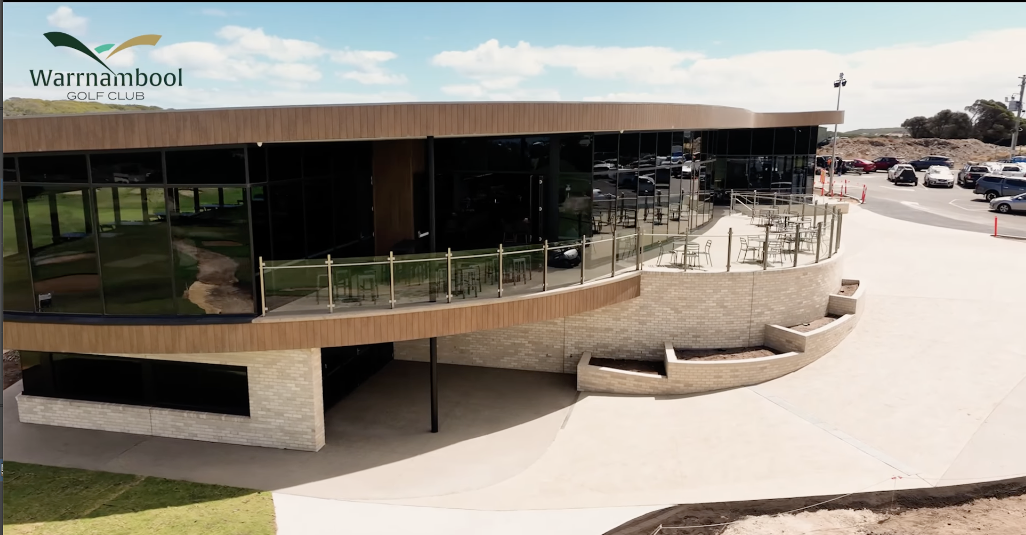 Exterior view of Warrnambool Golf Club building with glass windows, wooden and brick facade, outdoor seating area, parking lot, and cloudy sky.
