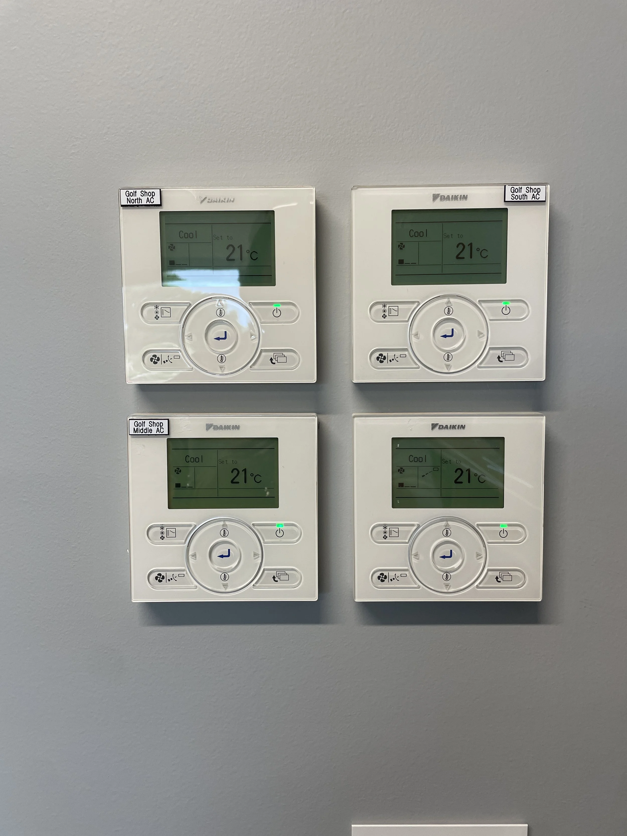 Four digital thermostats set at 21°C, mounted on a light gray wall, labeled for golf shop north, south, middle, and south AC.