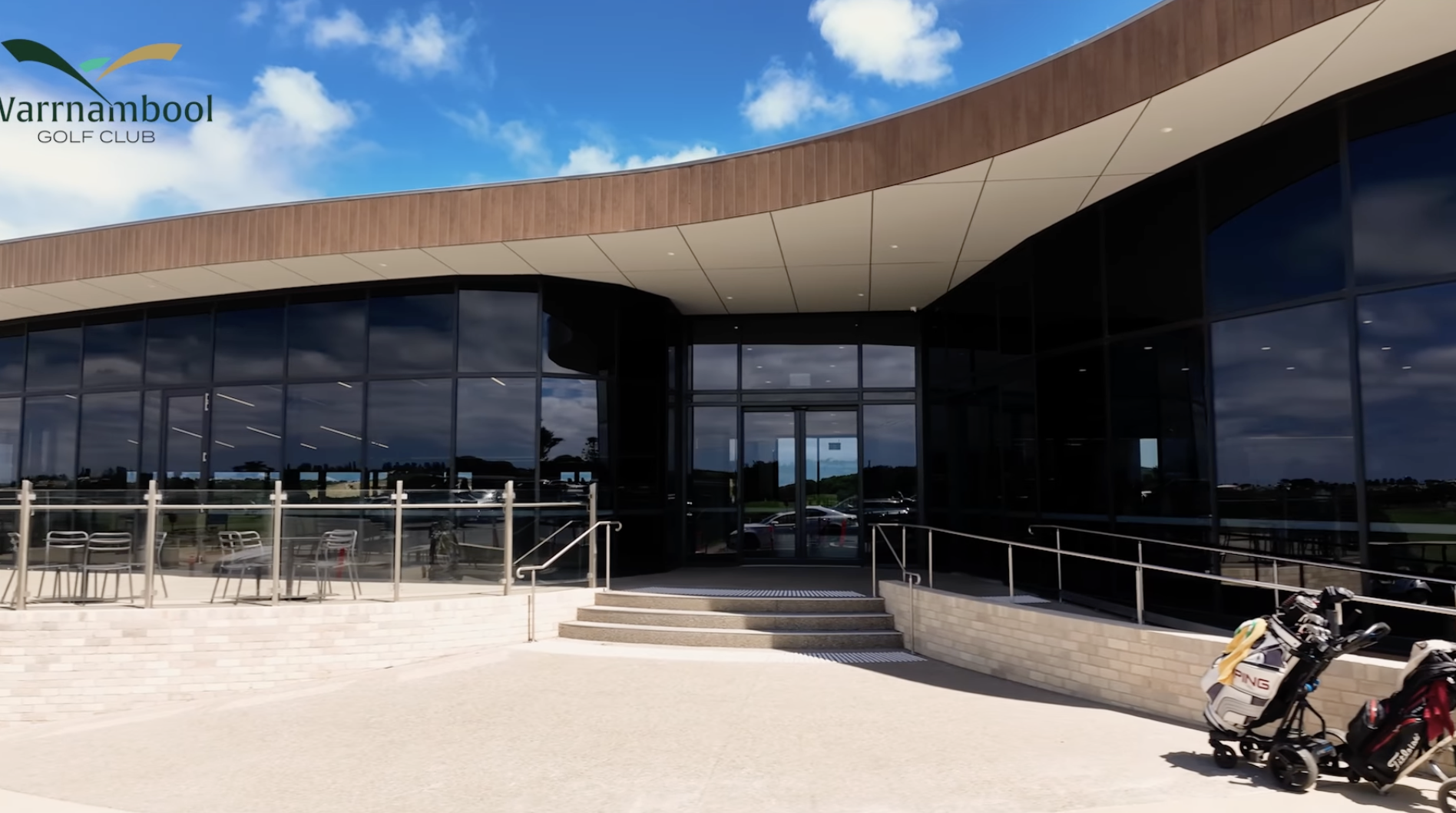 Facade of Warrnambool Golf Club with large glass windows, entrance stairs, golf bag with clubs, and blue sky with clouds.