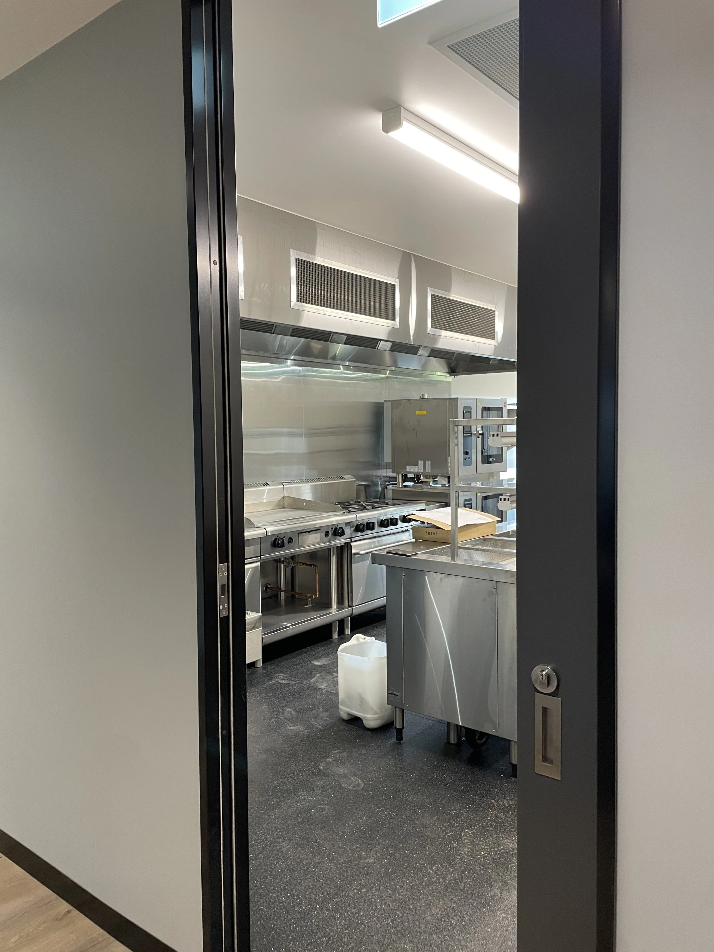 View into a stainless steel commercial kitchen seen through an open sliding door, with a stovetop, oven, and cabinets visible.