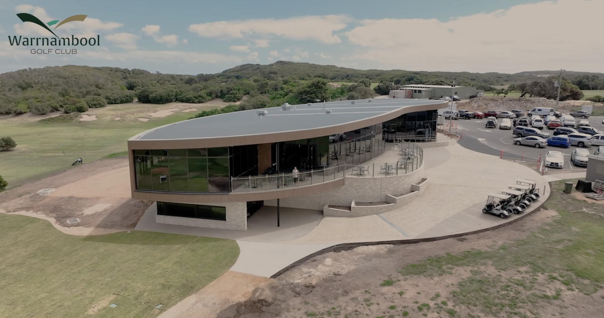 A modern golf club building with glass walls and a curved roof, surrounded by a golf course and parking lot, at Warrnambool Golf Club.