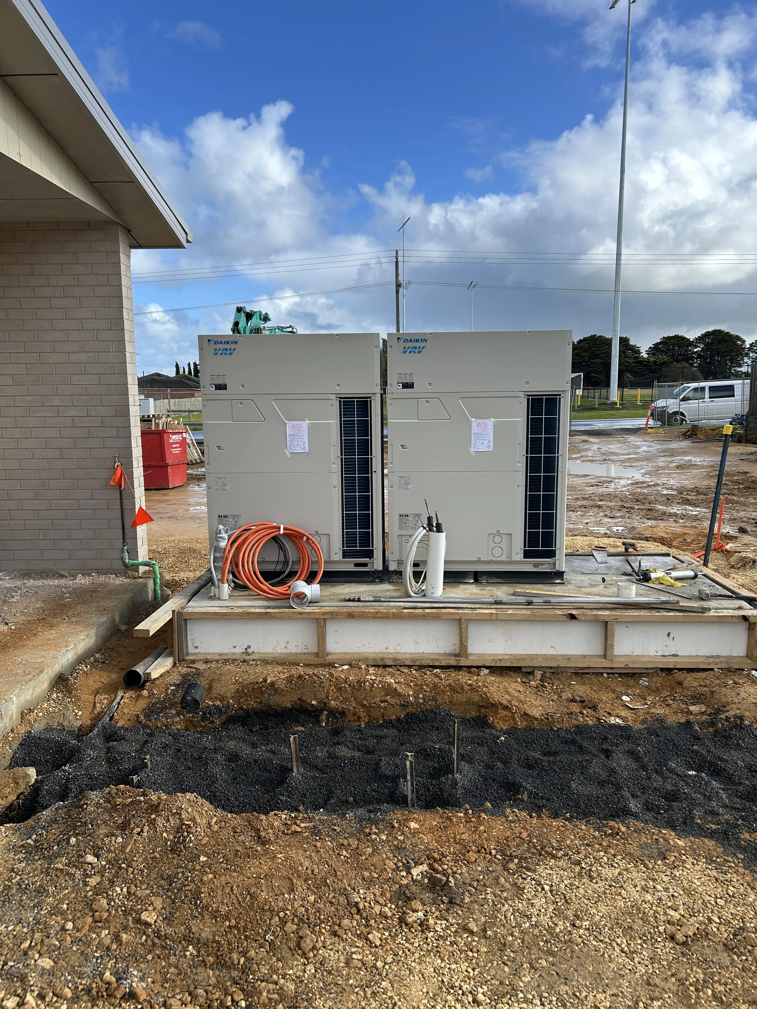Construction site with two large HVAC units installed outside a building, with assorted construction tools and piping, and a cloudy sky above.