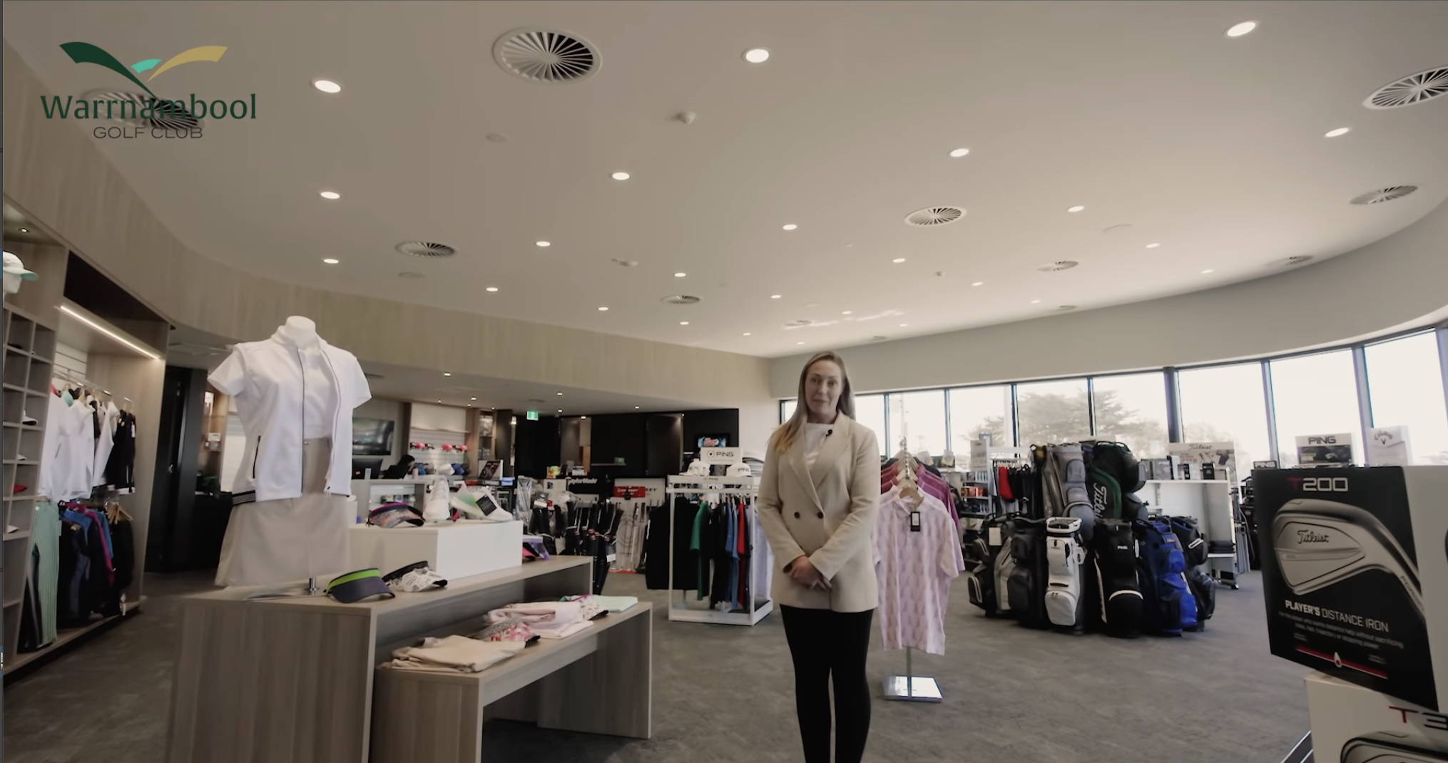 Inside a golf pro shop with clothing, golf bags, and equipment on display, and a woman in a beige coat standing near a clothing rack.