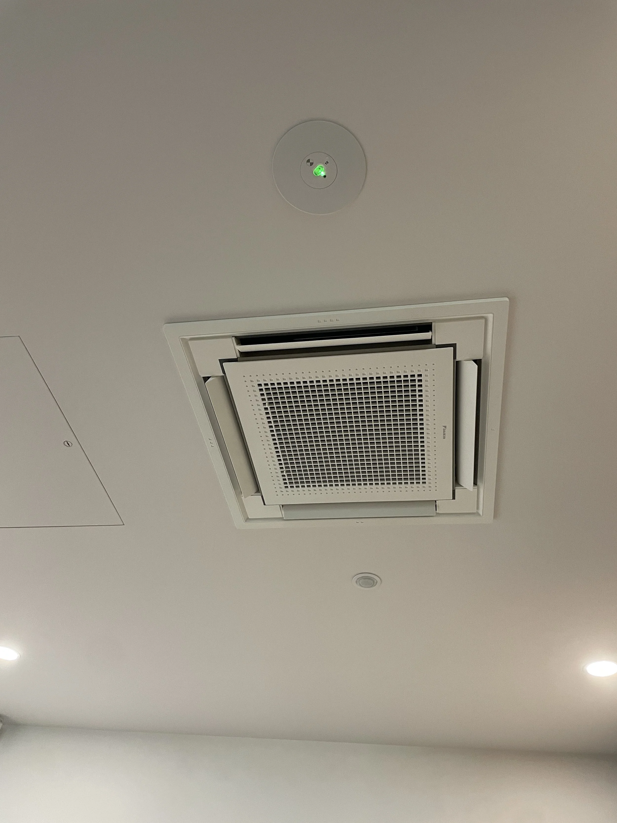 Ceiling-mounted air conditioning vent and smoke detector with green indicator light on a white ceiling.