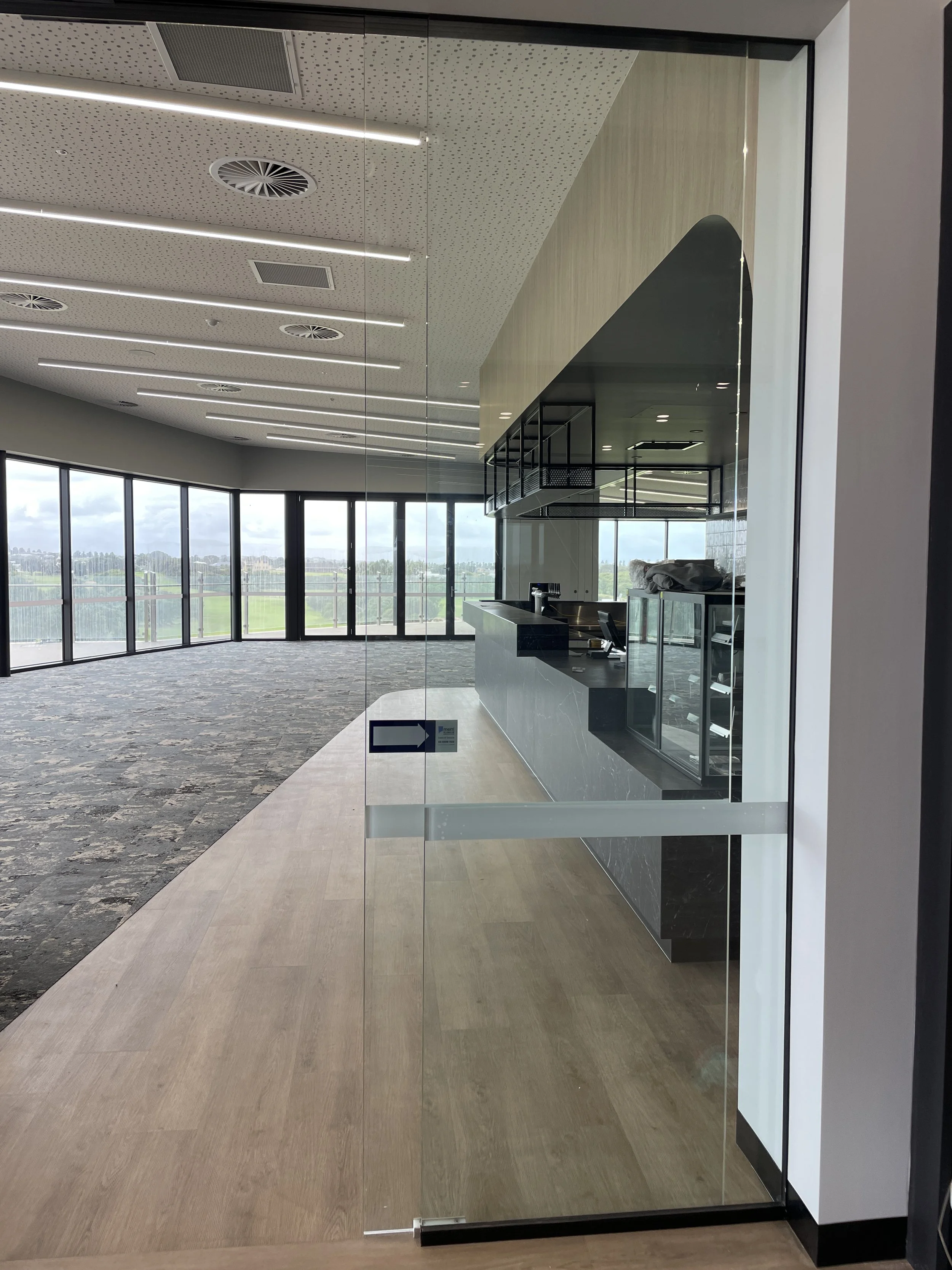 Empty modern lounge or cafe area with large glass windows, dark counter, and open view of a green landscape outside.