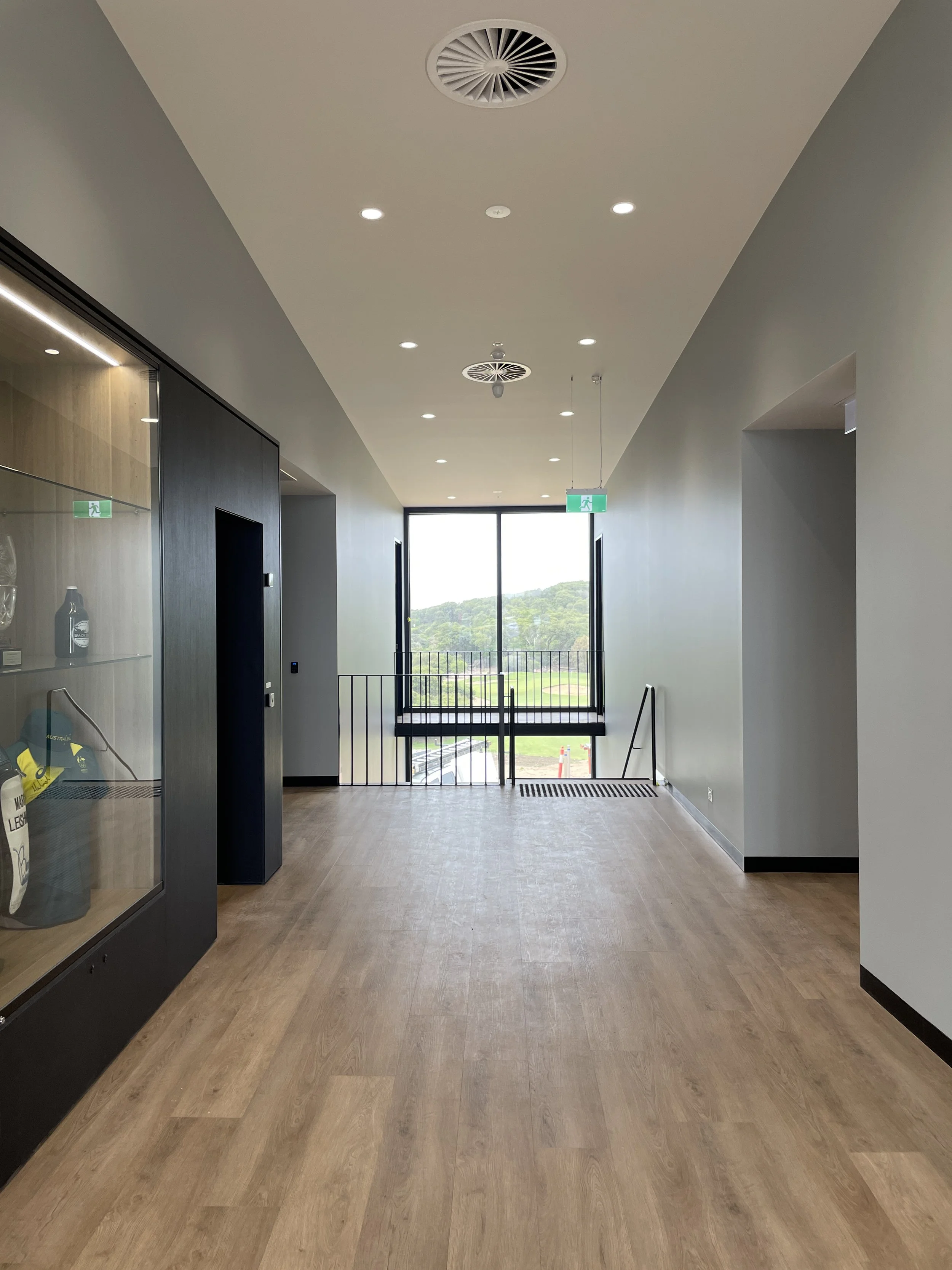 Empty modern hallway with wooden flooring, large window at the end, glass cabinet on the left, and exits with green emergency signs.