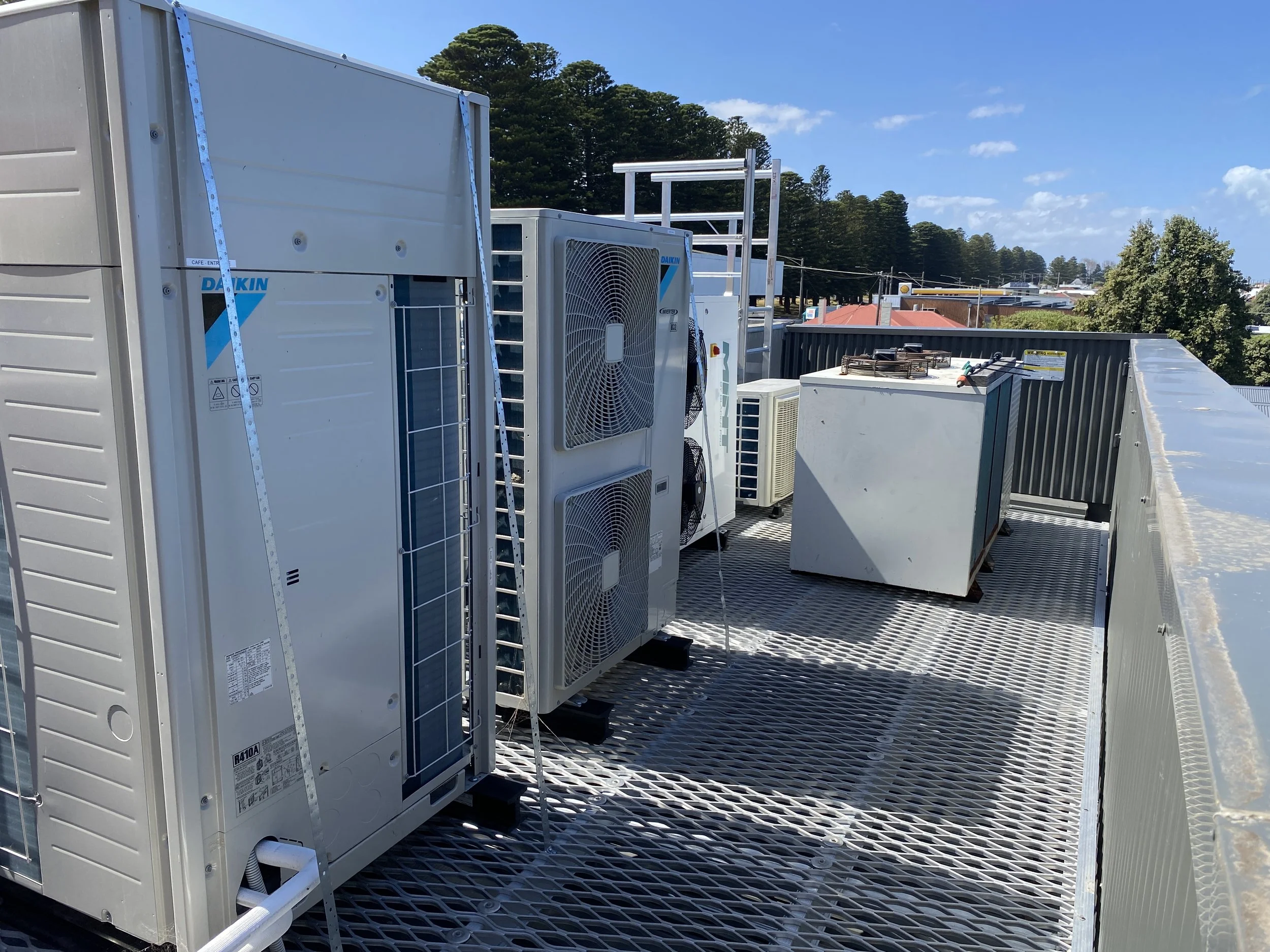 Rooftop with HVAC equipment including units, fans, and pipes, under a blue sky with some clouds, surrounded by a safety rail, with trees and buildings in the background.