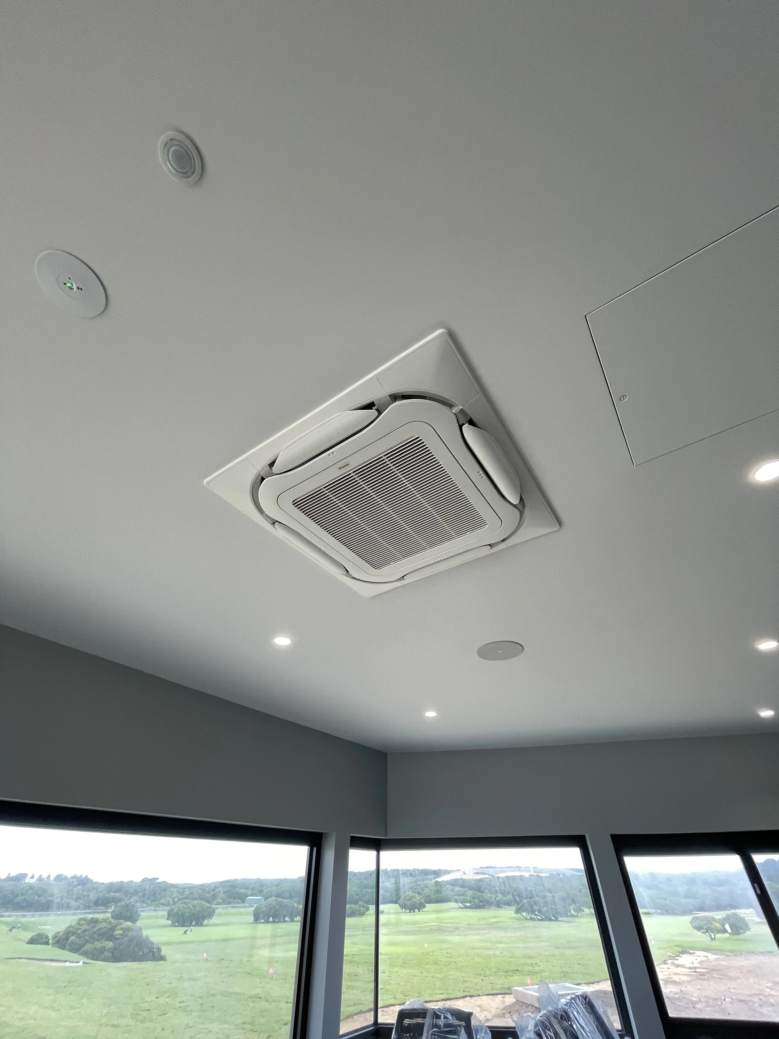 Ceiling with HVAC vent and recessed lighting in a room with large windows overlooking a green landscape.