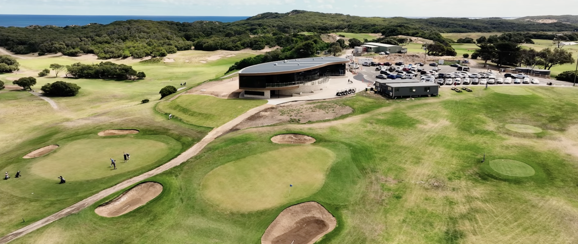 Aerial view of a golf course with sand traps, putting greens, and walking paths, adjacent to a modern clubhouse with a parking lot filled with cars, surrounded by trees and rolling hills, with the ocean visible in the background, Warrnambool.