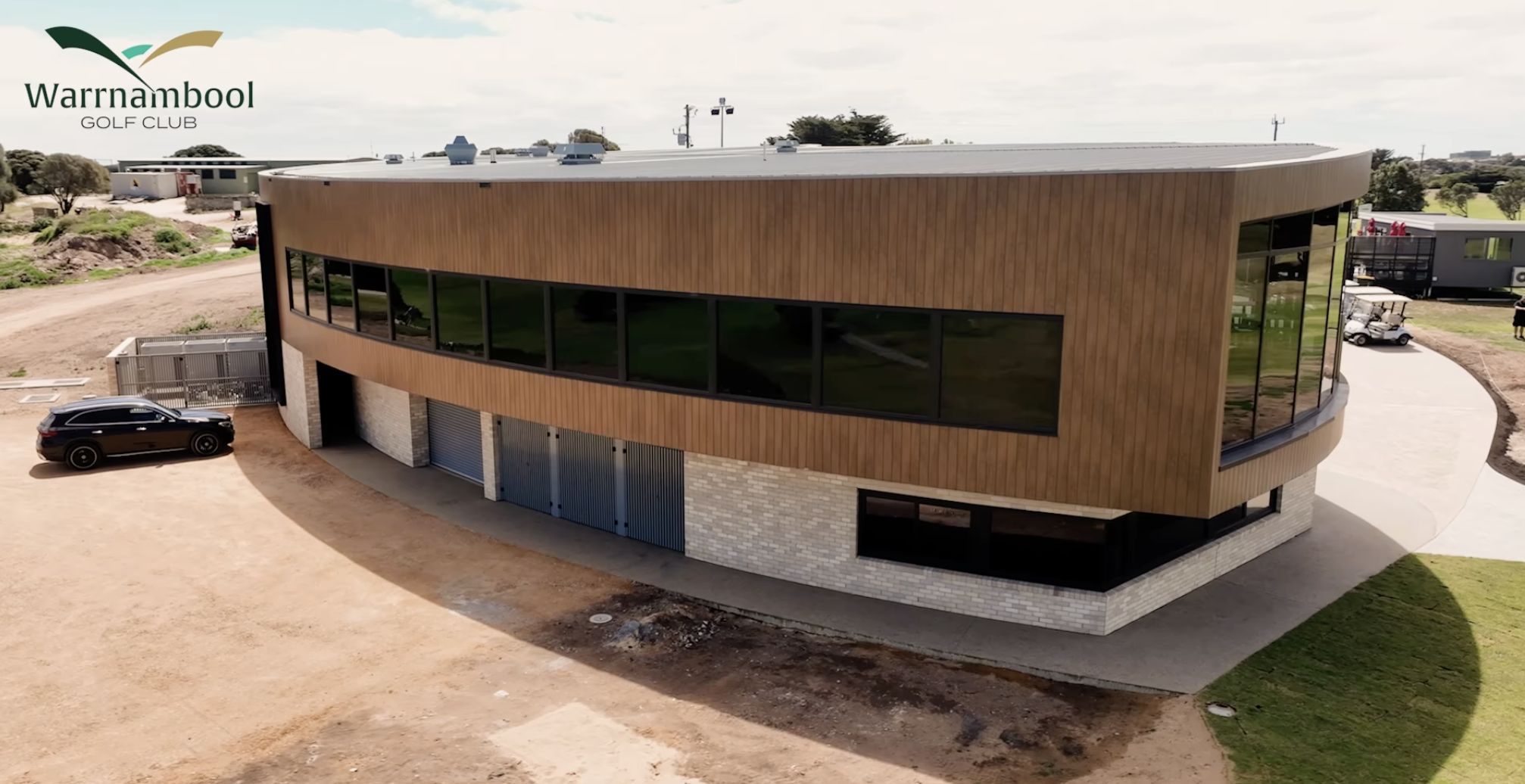 Modern building with curved design and large windows at Wynnambool Golf Club, with parked cars and surrounding landscape.
