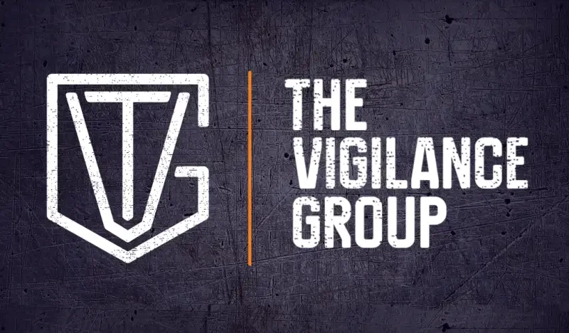 A logo with a shield outline on the left and the text 'The Vigilance Group' on the right, separated by an orange line, set against a dark, scratched background.