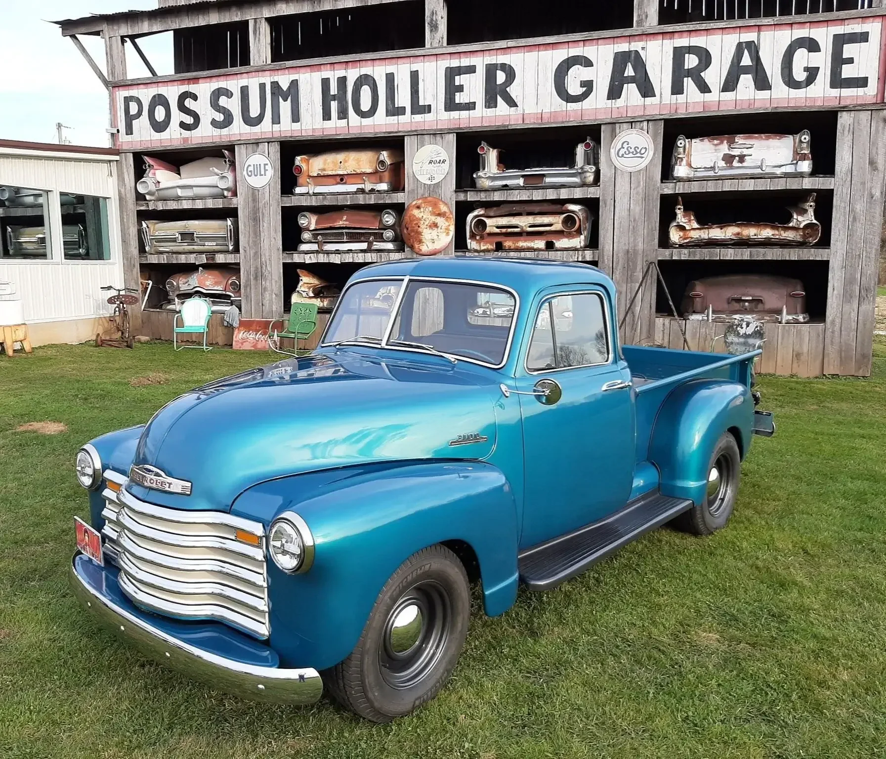 Vintage blue Chevrolet pickup truck parked on a grassy lawn with a wooden wall behind it displaying rusted car parts and signs, including 'Possum Holler Garage'.