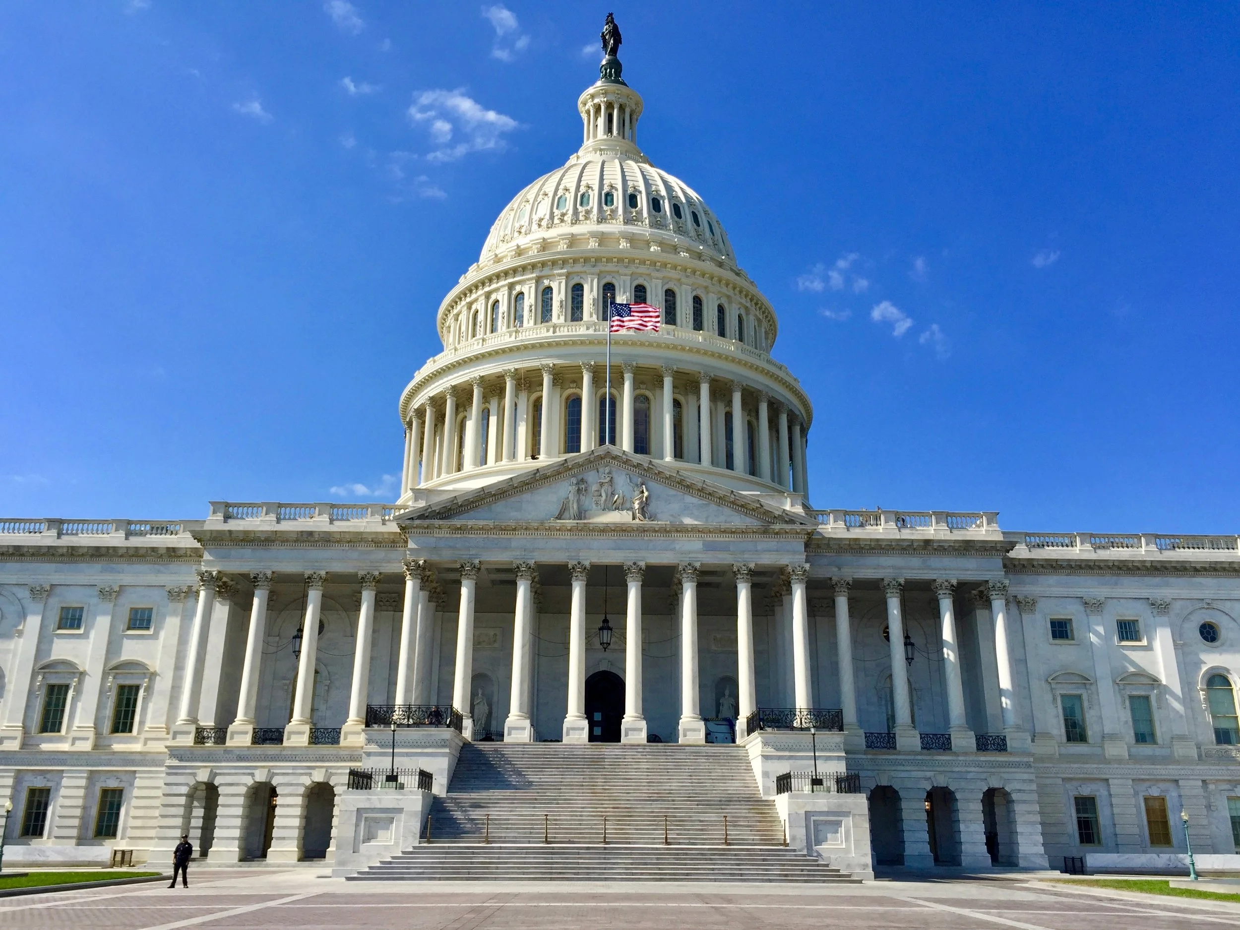The United States Capitol building with a clear blue sky, American flag, and a few people in front.