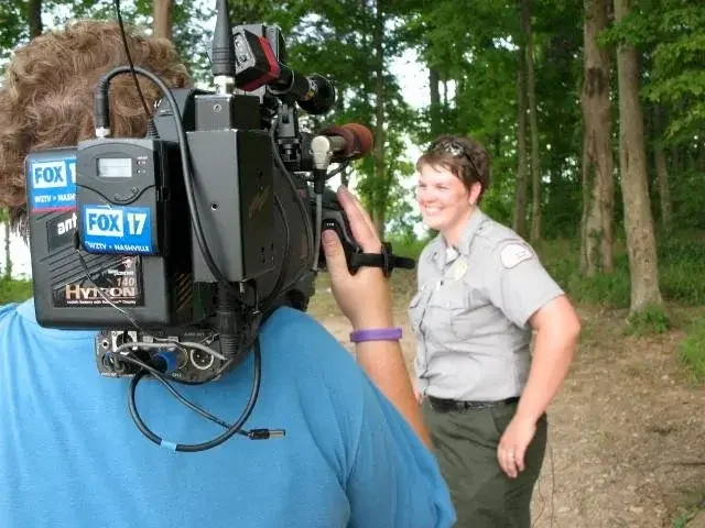 A TV crew filming a female U.S. Corps of Engineers ranger outdoors with trees in the background.