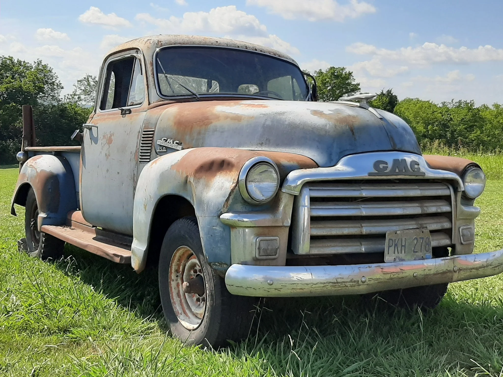 An old, rusty GMC pickup truck with peeling paint, parked on green grass under a blue sky with scattered clouds.