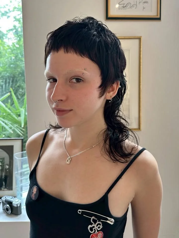A young woman with short black hair, piercings, and a septum nose ring, wearing a black spaghetti strap top with pins, standing indoors in front of framed artwork and a window with green foliage outside.