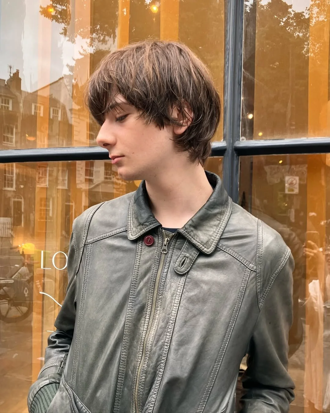 A young man with brown hair, wearing a gray leather jacket, stands in front of a large window with orange curtains inside, reflecting buildings and trees outside.