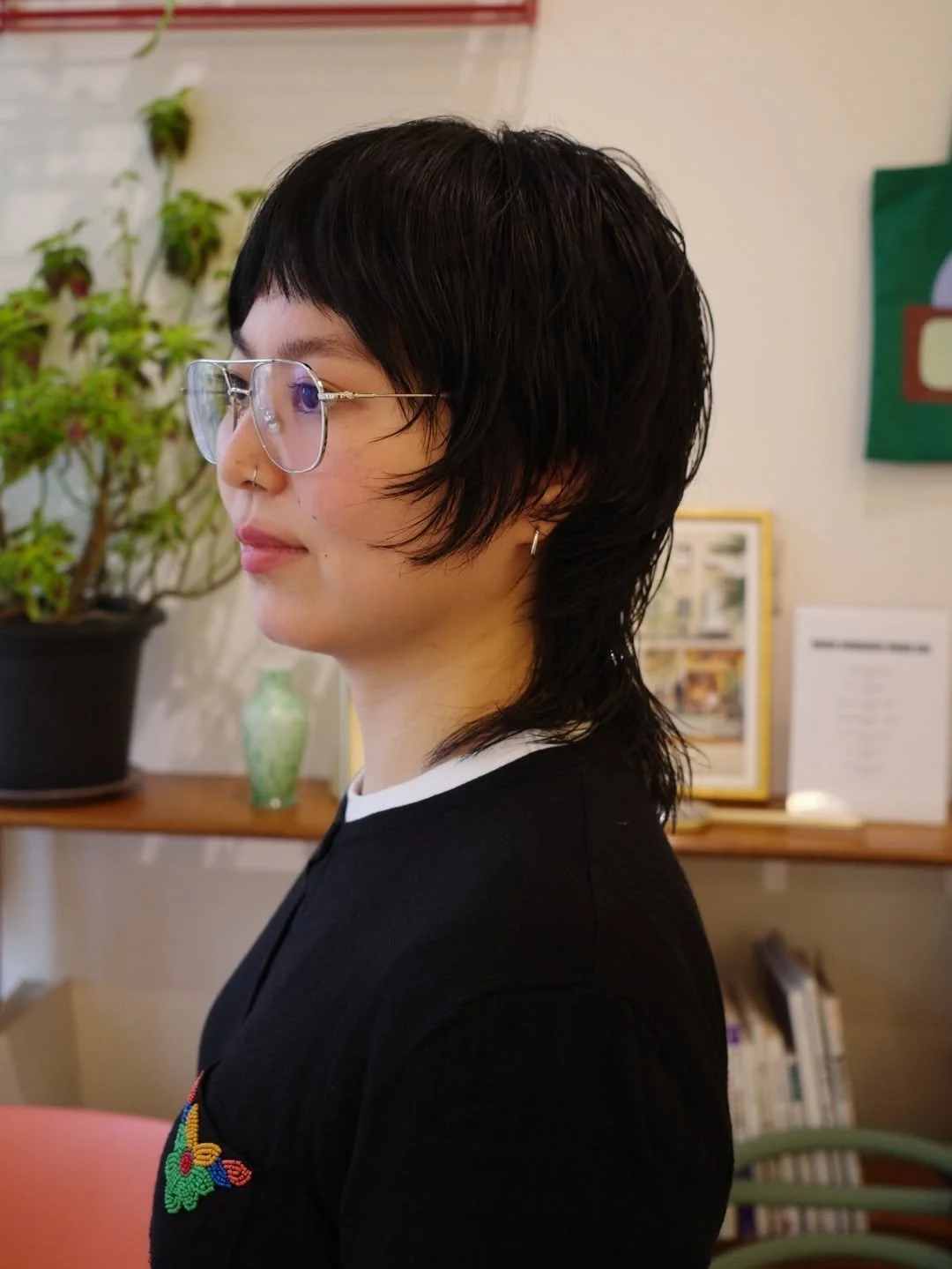 Side profile of a person with short black hair, glasses, and small earrings, indoors with a plant and framed pictures in the background.