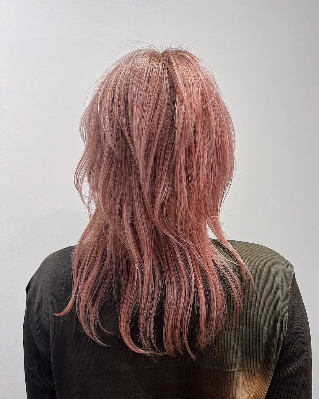 Back view of a person with wavy pastel pink hair wearing a dark green top.