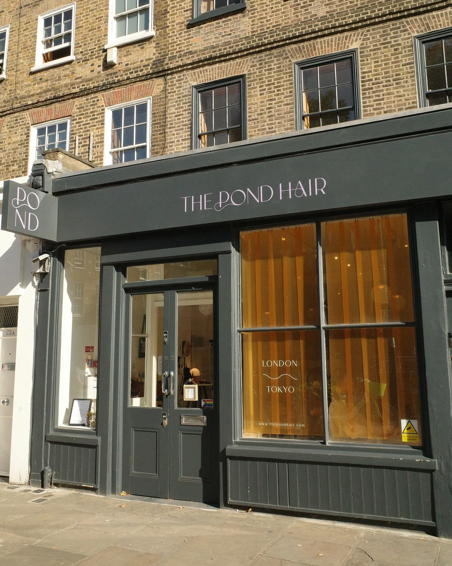 storefront of The Pond Hair salon with a gray facade, black door, and a large front window draped with yellow curtains. The shop sign reads "The Pond Hair", and on the window, it states "London" and "Tokyo".