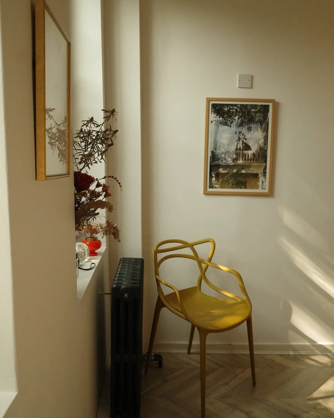 A cozy corner with a yellow modern chair, a window ledge with decorative items including a red vase and a silver spherical object, a black radiator, framed pictures on the wall, and sunlight casting shadows on the wall.