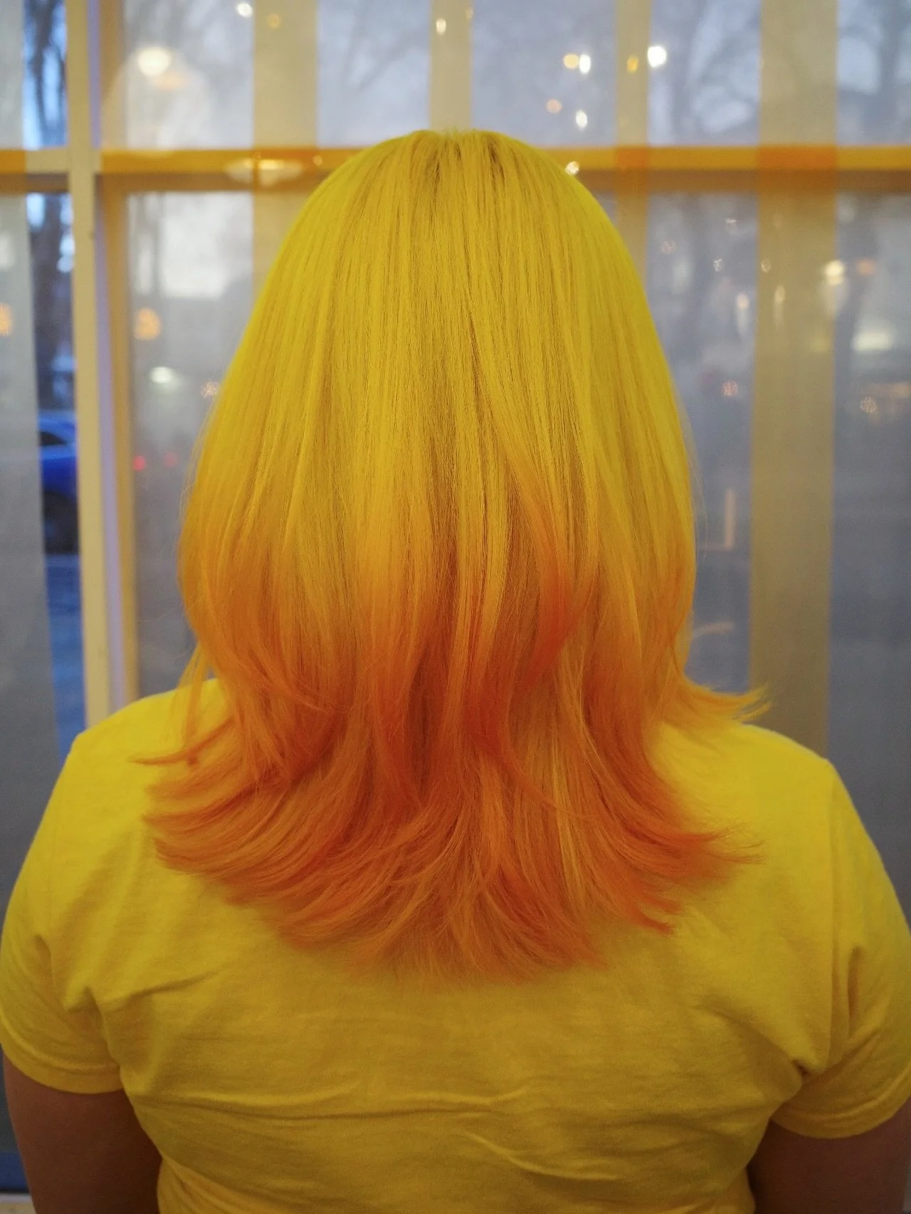 Back of a person with yellow and orange ombre hair wearing a yellow t-shirt, standing in front of a window.