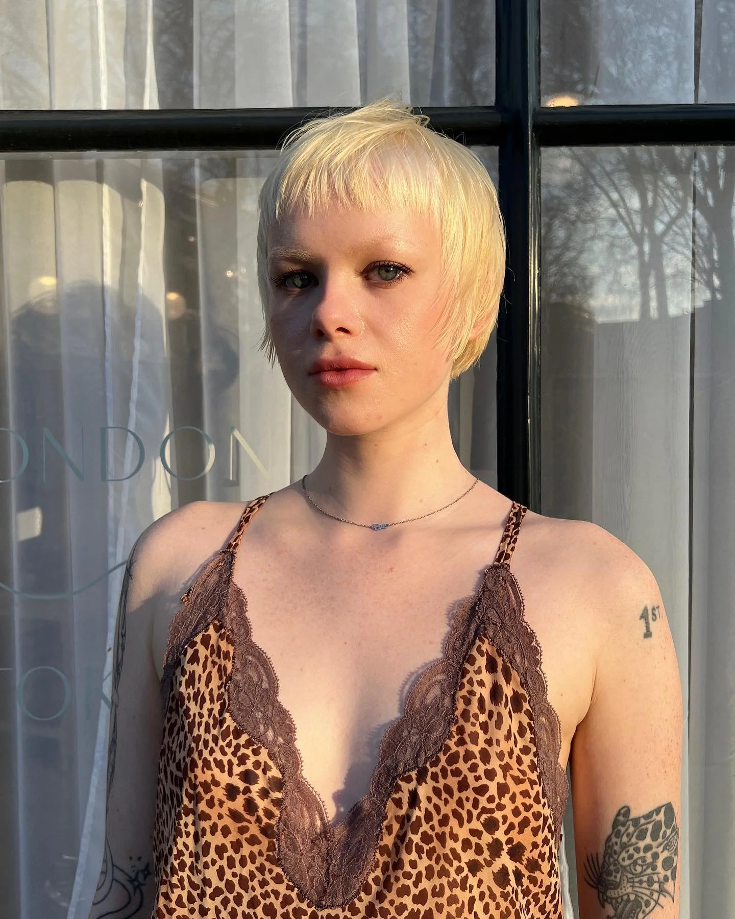 A woman with short blonde hair, wearing a leopard print dress with lace trim, standing in front of a window with curtains.