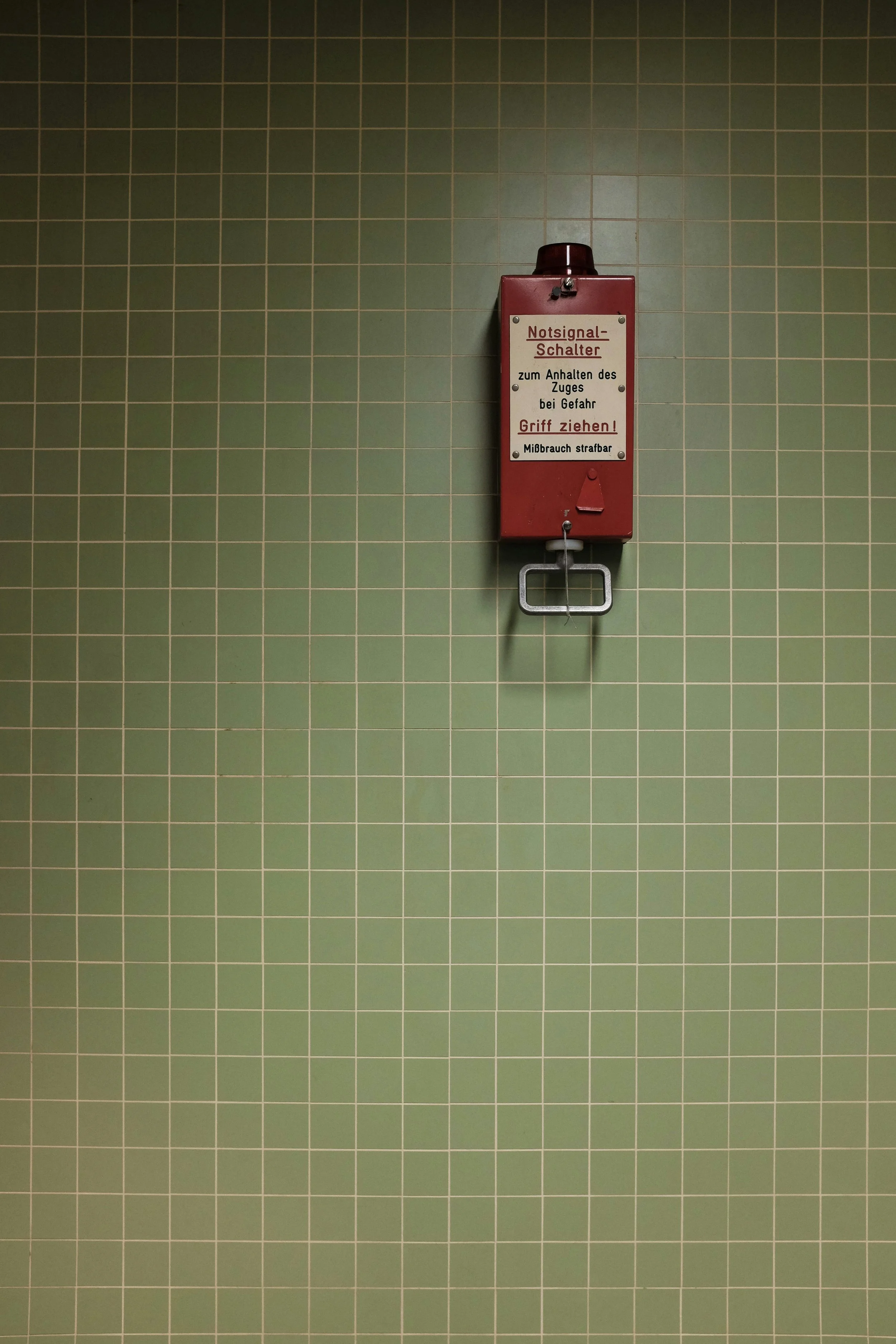 Red emergency signal switch box mounted on a green tiled wall.