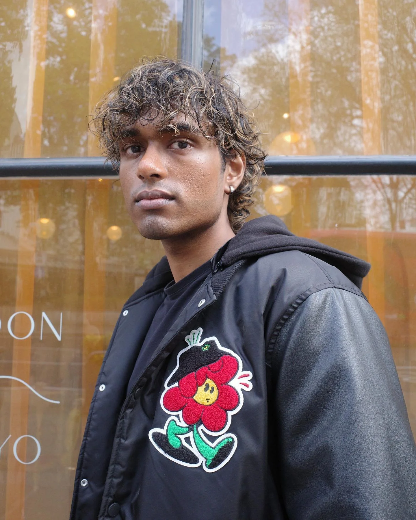 A young man with curly hair and earrings wears a black jacket with a colorful patch of a cartoon character on the front, standing outdoors in front of a glass window with yellow curtains and reflections of trees.