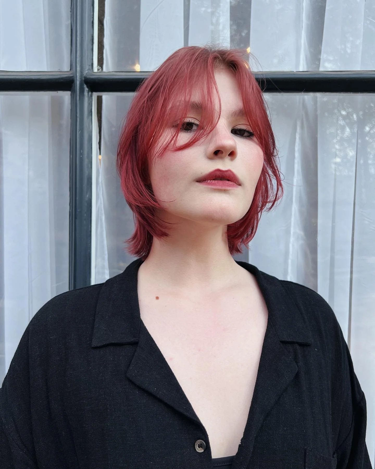 A young woman with shoulder-length red hair and fair skin looks at the camera, standing in front of a window with white curtains.
