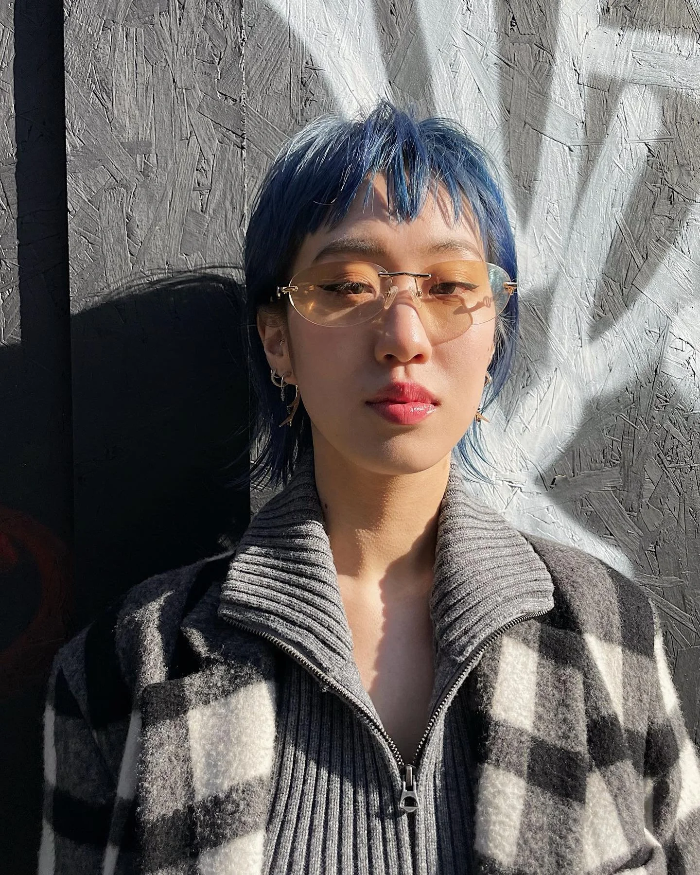 A woman with short blue hair, wearing sunglasses and earrings, posing against a textured gray wall with sunlight casting shadows.
